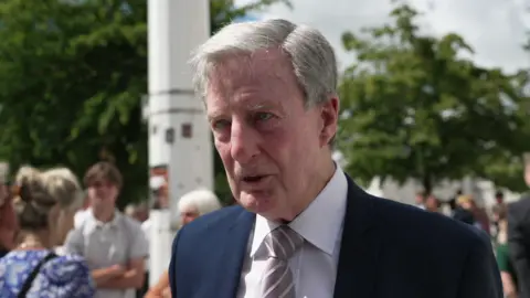 Pat Warne, son of Pte Bert Warne, being interviewed during the plaque unveiling event. He is wearing a suit and tie. Many people can be seen behind him.