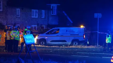 Supplied Police officers and National Highways staff stand beside a National Highways van near a road junction at night. Blue emergency lights are visible across the scene and debris can be seen on the carriageway.