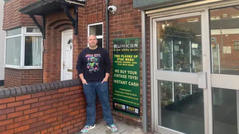 Simon Thake/BBC A middle-aged bald man in jeans and a black sweater stands with his hands in his pockets outside a red brick building next to a large green sign.