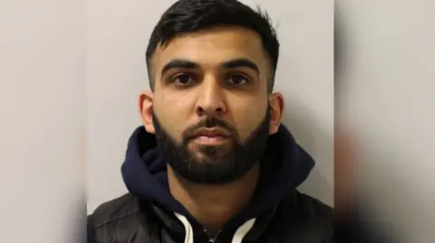 Met Police Man with dark beard and hair looks directly at the camera for a mugshot. He wears a dark blue hoodie and black gilet.