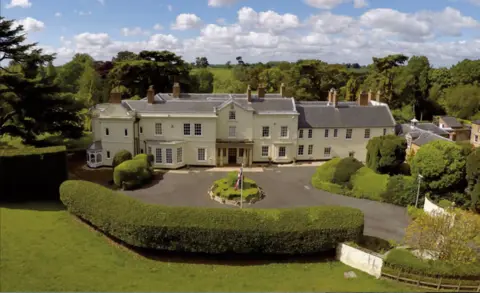 Moulton Grange is a cream coloured building with a grey slate roof and brick chimneys. This view shows the front façade, including an entrance porch with two columns on either side, a bay window to the left of the porch and multiple lattice pane windows. The approach to the property is a wide driveway surrounded by hedges with a circular bush in the centre of the drive and a union flag on a white pole.