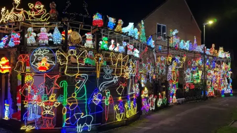 Sue Ryder A house is surrounded by a large cage-like structure which is covered in lit-up Christmas decorations of stars, the Nativity, gingerbread men and women, bells, angels, and more. It is dark and a street light is on.