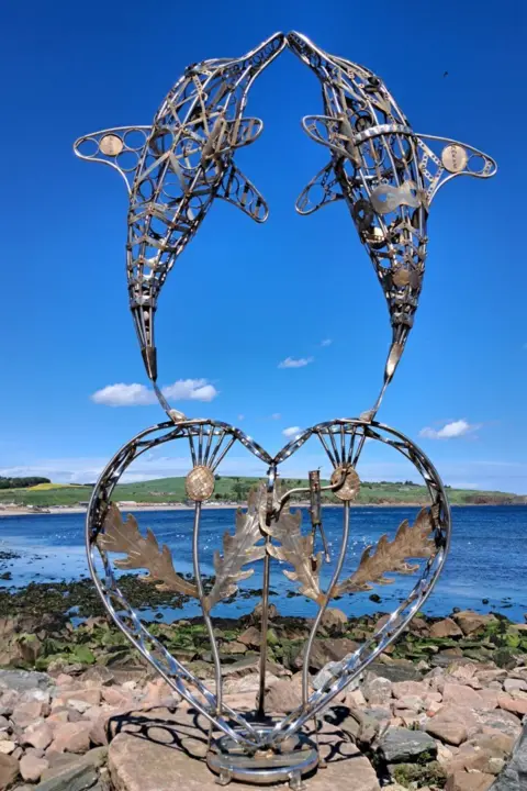 Angela Pearson A metal sculpture of two dolphins on the shore in Stonehaven. It stands on a rock with pebbles on the beach and the sea in the background. The sky is clear with few clouds. 