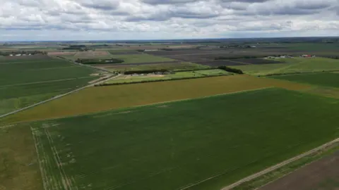 Steve Hubbard/BBC Aerial image of a patchwork of green and brown ploughed fields, with some dry patches and various buildings in the background. It is a cloudy day.