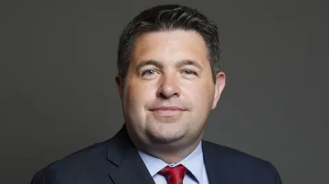 UK Parliament MP posing for official House of Commons photo against a plain, dark background