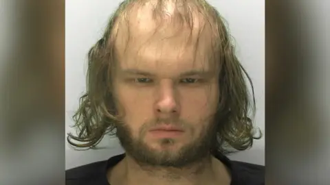 Victor scowls at the camera in a police custody picture. He has a short scruffy beard and thin scraggly hair which falls over his face. 