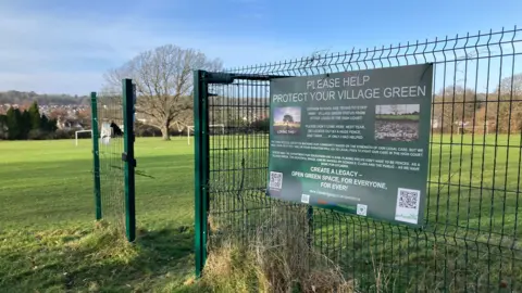 BBC A fence in the middle of a lush green field pictured on a sunny day has a sign that reads "Please help protect your village green".