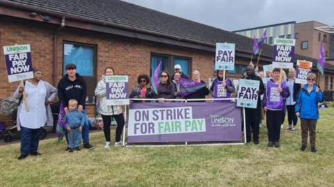 Staff with placards "on strike for fair pay" on grass outside the hospital