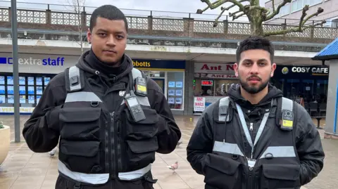 Two men in black security guard outfits. Kitted out with a body worn camera and radio. They are posing for a picture on a precinct square in Basildon town centre.
