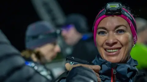 Mid Cheshire Hospitals NHS Foundation Trust A woman wearing a dark blue coat as well as a pink bandana and a headtorch is standing outdoors at night among a group of other people. She is smiling while looking at the camera.