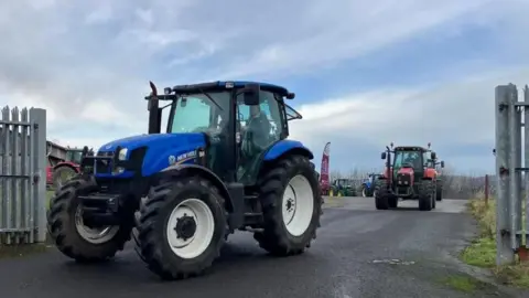 A blue tractor leads the rally of tractors. A red tractor can be seen following it and others behind that.