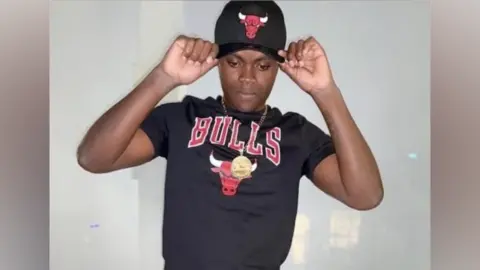 Khadeem Parris, a teenager, poses in front of the camera. He wears a Chicago Bulls branded black T-Shirt and black baseball cap. The T-Shirt is printed with the word BULLS in red, and the red face of a bull. The cap also has the red face of a bull printed on it. Khadeem holds the edges of the baseball cap with his finger tips. He wears a chain round his neck, and a white wall is behind him.