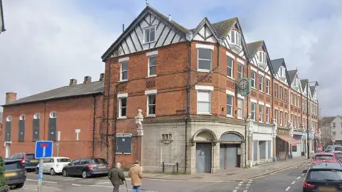 Google The outside of Weybridge Hall which is a three storey red brick building which extends out the back to at two floor height. It has pitched roofs which are white and black at the top and is on the corner of two roads.