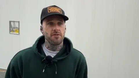 Michael Broomhead/LDRS A man with tattoos on his face and neck. He is wearing a dark green hooded jumper and a baseball cap.