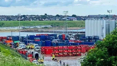 Emergency services at an industrial base, with sea water and then houses in the distance.