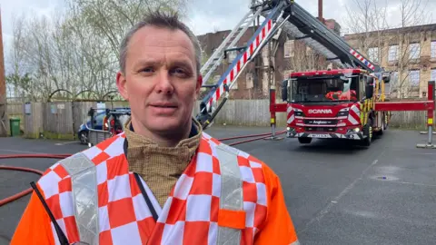 A man in an orange and white check jacket with high-vis stripes either side of the collar. He stands in front of a fire engine with ladder extended. A large mill building can be seen behind.