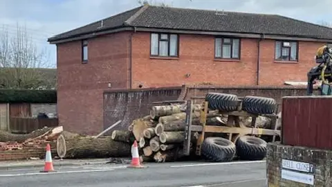 An overturned tractor trailer which has shed its loads of logs on the A38 West Huntspill in Somerset. Damage can be seen to the wall that the tractor crashed into. There are three traffic cones cordoning off the area.