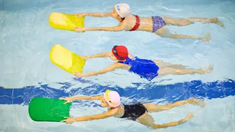 An aerial view of three children swimming in a pool and holding floats out in front of them. Two have pink swimming hats, while another has a red one