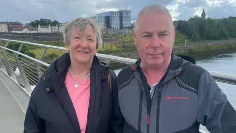 a man and woman stand side by side on a bridge, a river is behind them, and a number of buildings can be seen in the background
