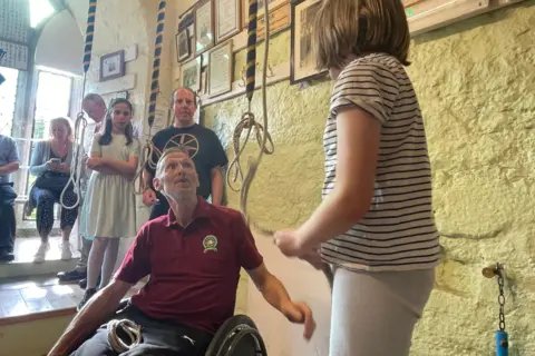 BBC Mike is in a wheelchair and looks up at a girl who is pulling a rope for a bell. They are in a bell towers surrounded by the other ringers.