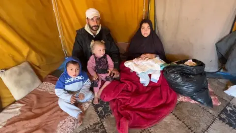 A man, a woman and two small children are sitting in a tent with a bin bag