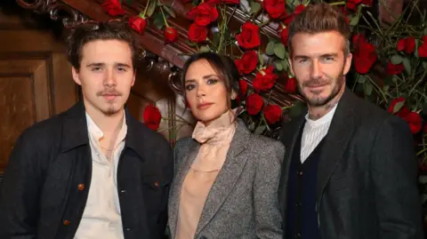 Getty Images Brooklyn Beckham (left) with Victoria Beckham (centre) and David Beckham (right) they all pose at the camera. They stand in front of a staircase decorated with red roses. 