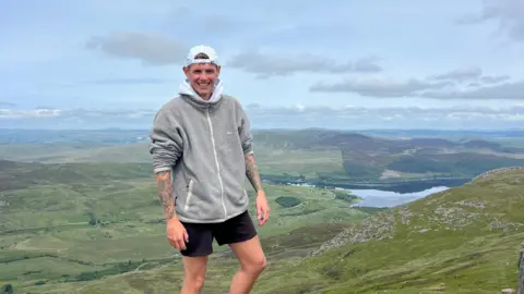 Ed Shirt Ed Shirt wearing a hoodie and shorts standing on a mountain with a view behind him