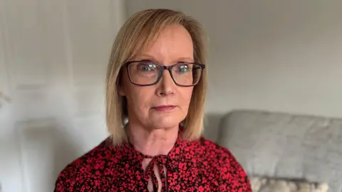 A woman with shoulder length straight blonde hair looks at the camera. She's wearing a black blouse with red flowers