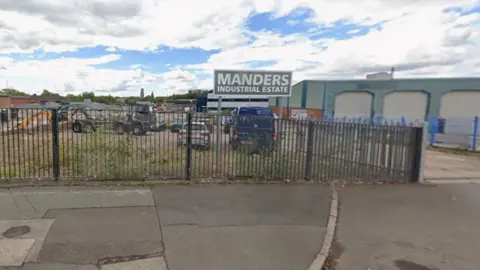 A pavement is in the foreground, with the words Manders industrial estate on a sign behind a fence. Vehicles and a building are behind the sign.