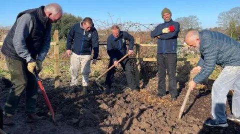 A group of five men wearing outdoor clothing are gathered around a mound of dirt. Some of the men are digging using long shovels and others are standing overseeing the work.