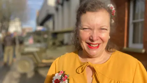 BBC May Rose wearing a yellow top with a pink and white flower brooch 