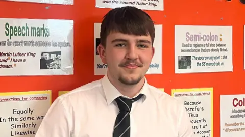 Tomos is staring at the camera wearing a white shire and blue tie. Stood in front of an orange notice board with white papers on it