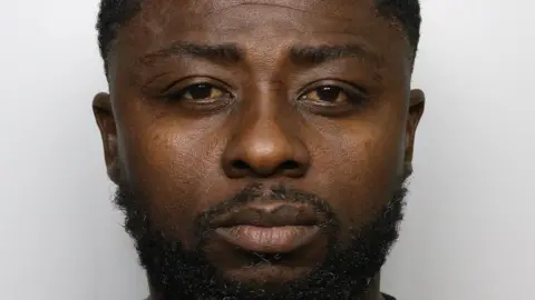 A mugshot of Boachie, photographed from the front against a plain, light‑coloured background. He has short, closely trimmed hair and a beard. He is wearing a dark T‑shirt.