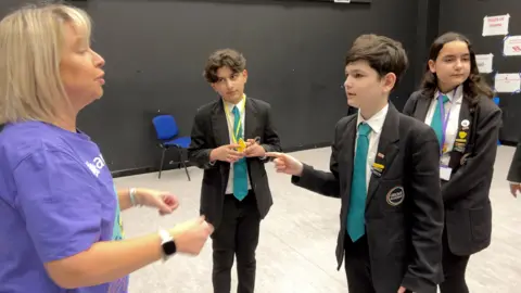 BBC A blonde haired lady wearing a purple t-shirt talks to a teenage boy wearing a blazer and school tie in a drama studio.