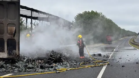 A firefighter wearing a blue and yellow fire suit, holding a yellow hosepipe, putting out a fire on the road. Debris and smoke can be seen on the road.