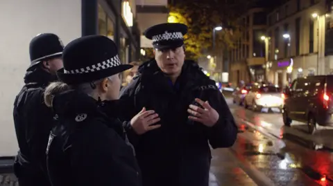 Avon and Somerset Police Chief Constable Crew in uniform in the foreground speaking to Neighbourhood Inspector Dudley Bond in Bath. They are on a busy street with lots of cars driving down it at night. Everyone is wearing full police uniform, including caps and hats.  