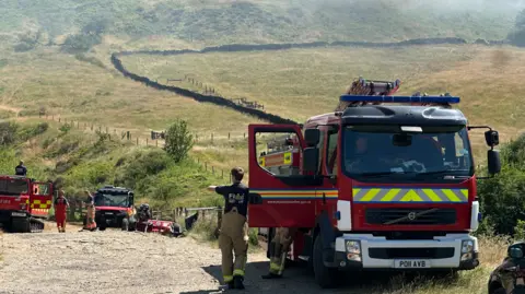 Three firefighters are standing or walking near to fire engines parked on a dirt track. Smoke can be seen on the moors in the distance.