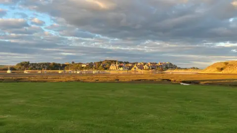 The village of Alnmouth is bathed in late evening sunshine. It sits on a sandy outcrop with lots of houses painted different colours and a sandy bay stretching around it 
