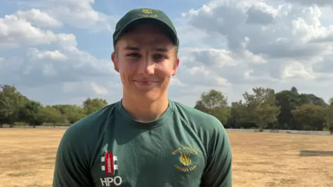 Young man with a green cap on and a green long sleeve top. He is smiling. He is stood on the edge of a cricket field and the grass is parched and sandy coloured. 