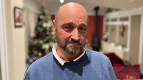Gary Gilding, a middle-aged white man wearing a blue sweater revealing white collars underneath. He is standing in a living room which is blurred in the image but shows a Christmas tree and a log wood burner. 