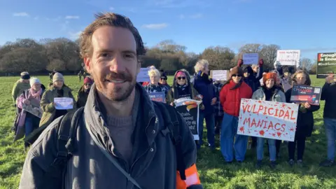Jake Zuckerman/ BBC Laurence Bradley stood in front of a group of protesters holding up signs. He has short brown hair and facial hair and is wearing a black coat. The sky is blue.