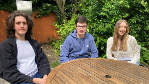 Left to right: Charlie, James and Lucie, sitting around a wooden table in a garden