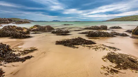 Dennis Clarke Seaweed-covered rocks dot a shallow bay with pale sand and turquoise-green water. The long-exposure photograph creates a smooth, flowing effect in the water beneath dramatic blue-grey clouds and distant low hills.