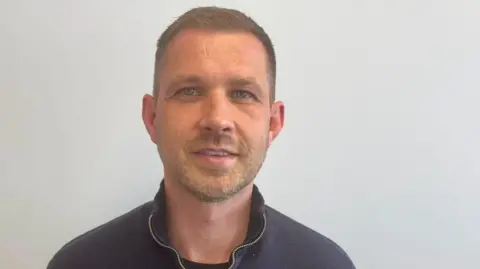 A man with short hair and stubble, wearing a blue jumper, is standing in front of a white background and smiling at the camera.