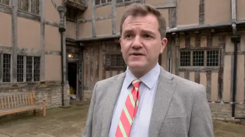 Luke Purser wearing a grey blazer and a red and white tie. He has brown hair and is standing in front of Hall's Croft. Hall's Croft is a brown and black building.