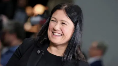 Head and shoulders shot of Nandy, who wears a black t-shirt under a black jacket, and a large red Labour rosette on her lapel. She wears her long black hair loose over her shoulders and is smiling broadly.