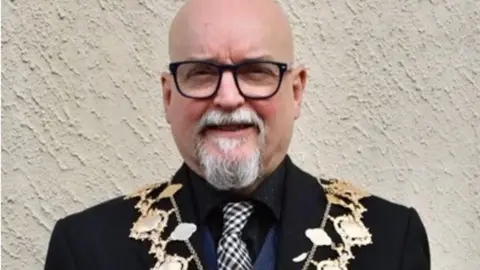 An elderly bald man with black-framed glasses and a white goatee beard smiles at the camera. He is wearing a dark suit, a black shirt and a checkered tie, as well as a chain of office.