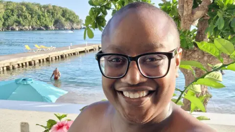 Simone Boothe, wearing glasses, smiles on holiday with a beach in the background.