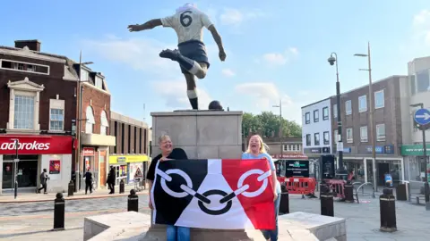 Two women are standing in front of a statue in Bilston town centre. They are holding the flag of the Black Country. The flag is black, white and red with chain representing the region's manufacturing heritage.