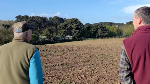 Two men face a brown ploughed field 